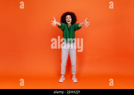 Come to my arms. Full length of friendly young adult woman with Afro hairstyle wearing green casual style sweater spreading hands, inviting, embracing. Indoor studio shot isolated on orange background Stock Photo