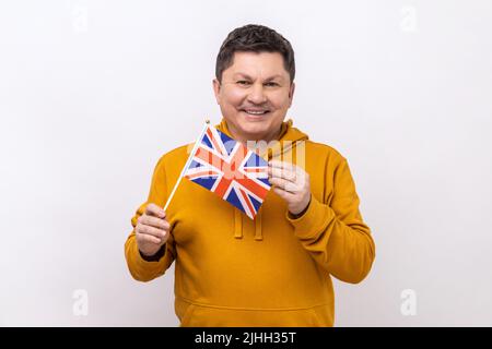 Portrait of smiling delighted handsome middle aged man holding flag of United Kingdom of Great Britain, wearing urban style hoodie. Indoor studio shot isolated on white background. Stock Photo