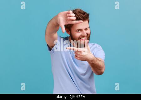 Portrait of curious bearded man gesturing picture frame with hands, looking through fingers and focusing on interesting moment, taking photo. Indoor studio shot isolated on blue background. Stock Photo