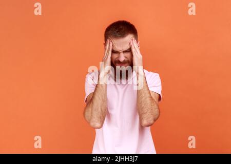 Portrait of unhealthy sick young adult bearded man massaging temples, suffering from headache, migraine, wearing pink T-shirt. Indoor studio shot isolated on orange background. Stock Photo