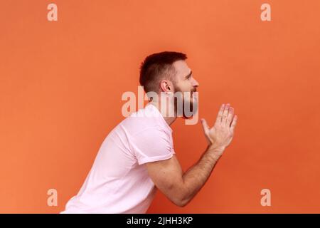 Side view of bearded man folding hands in prayer, closing eyes and talking to god, asking for help, expressing gratitude, wearing pink T-shirt. Indoor studio shot isolated on orange background. Stock Photo