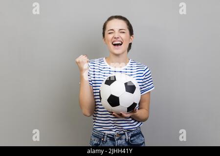 Portrait of woman wearing striped T-shirt screaming holding soccer ball, celebrating victory of favourite football team on championship. Indoor studio shot isolated on gray background. Stock Photo