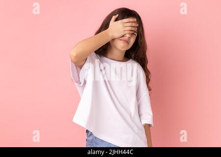 Don't want to look at this. Portrait of little girl wearing white T-shirt covering eyes with hand, feeling shamed and scared to watch. Indoor studio shot isolated on pink background. Stock Photo