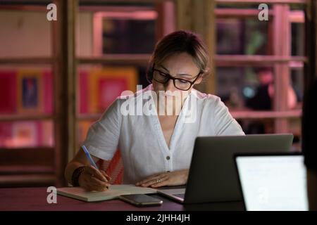 Young focused concentrated female student wearing spectacles studying in cozy cafe using laptop Stock Photo