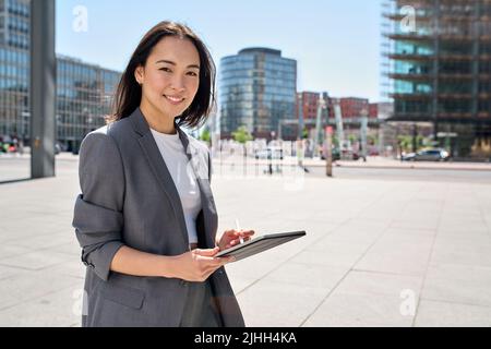 Young happy Asian business woman standing on city street holding digital tablet. Stock Photo
