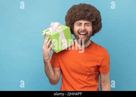 Portrait of extremely happy man with Afro hairstyle wearing orange T-shirt holding green present box and looking at camera with joyful expression. Indoor studio shot isolated on blue background. Stock Photo