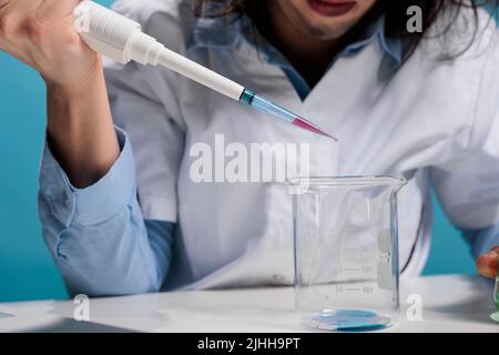 Close up portrait of wacky insane chemist using dropper to mix experimental chemical compounds after lab explosion. Foolish mad looking scientist with pipette doing experiments while sitting at desk. Stock Photo