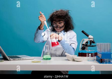 Mad scientist with wild appearance acting silly after lab explosion. Portrait of crazy amusing chemist acting funny and having dirty face and messy hair while sitting at desk and looking at camera Stock Photo