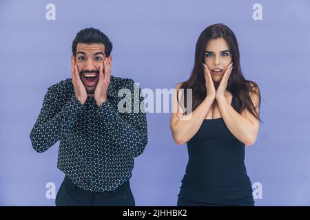 Surprised young heterosexual couple of beautiful young adults in black clothing looking at camera and touching their cheeks. Isolated on violet background. High quality photo Stock Photo