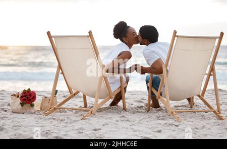 Happy african American couple spending a day at the sea together. Content boyfriend and girlfriend kissing while sitting on the beach. Caring husband Stock Photo