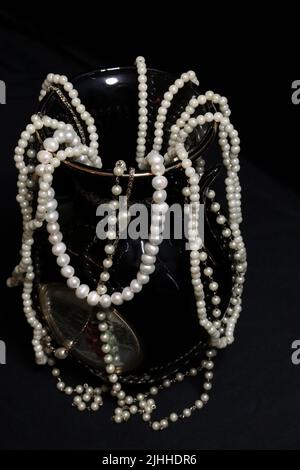 A pile of various pearl necklaces up close and on a neutral backdrop with add-ons like a vase, old hat, mask, and red/gold storage canisters. Stock Photo