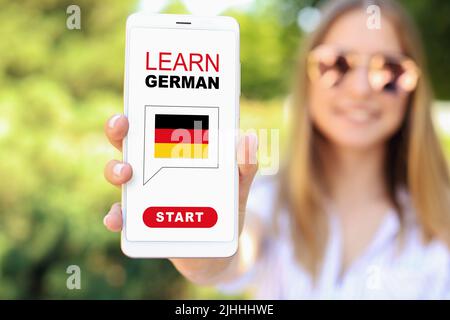 Woman holding mobile phone with text LEARN GERMAN on screen outdoors, closeup Stock Photo