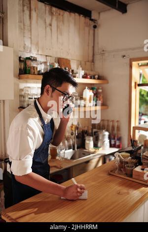Young barista accepting order from customer in phone and writing it on notepad Stock Photo