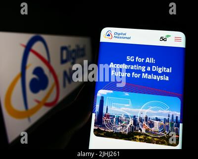 Person holding cellphone with webpage of Malaysian company Digital Nasional Berhad (DNB) on screen with logo. Focus on center of phone display. Stock Photo