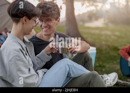 Cheerful couple in love sitting on grass in camping, laughing and cheers with cups of tea. Stock Photo