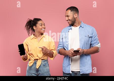 Positive black couple using smartphones, happy woman showing her gadget to boyfriend, sharing social media content Stock Photo