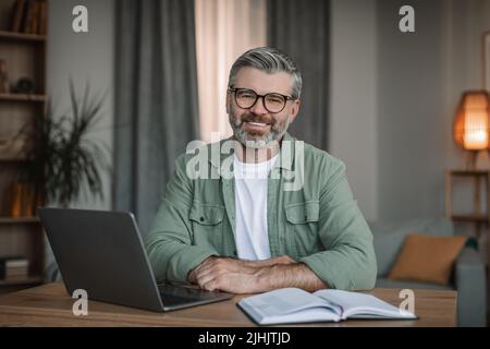 Glad mature caucasian male teacher with beard in glasses working with laptop, shooting video lesson in room Stock Photo