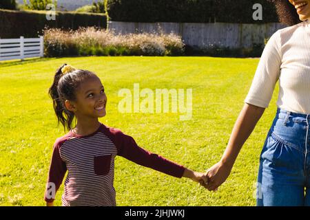 Midsection of biracial mother holding happy daughter's hands while standing on field in yard Stock Photo