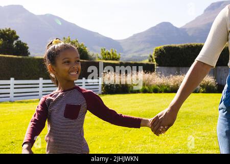Midsection of biracial mother holding smiling daughter's hands while standing on field in yard Stock Photo