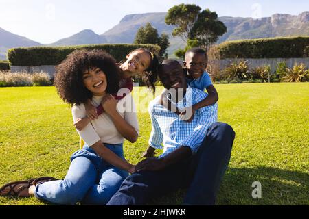 Portrait of happy multiracial children cuddling parents from behind sitting on grassy field in yard Stock Photo