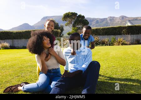 Happy multiracial children cuddling cheerful parents from behind sitting on grassy field in yard Stock Photo