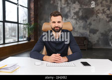 Portrait of handsome young businessman in smart casual wear sitting at the desk in loft office and looks at camera, male employee involved virtual meeting, webcam view Stock Photo