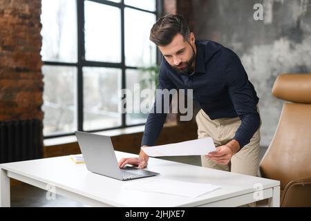 Busy businessman looking through the workpaper and using laptop in modern loft office, hurrying to finish report in time Stock Photo