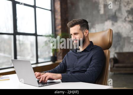 Concentrated determined young bearded office worker in business casual clothes, sitting at the desk, typing on laptop, businessman professional is working on project, writing email to client Stock Photo