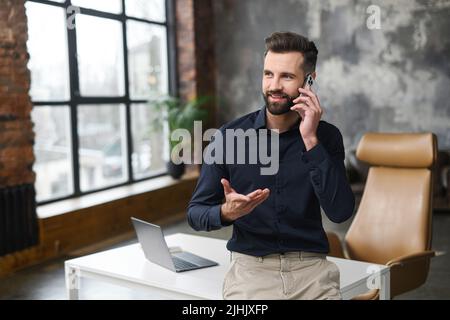 Proud and successful young businessman in smart casual wear talking on smartphone, male entrepreneur, start-up owner has phone conversation standing in the office Stock Photo