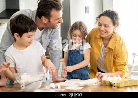Happy family of four cooking in the kitchen together, parents and two kids making dough together, preparing pie or cookie, have a fun on weekend Stock Photo