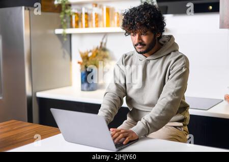 Clever Indian freelancer using laptop for remote work from home. Handsome bearded guy web surfing, sending and answering emails, developing software while standing. Intelligent man working remotely Stock Photo