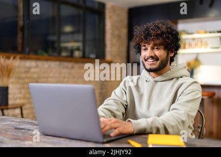 Happy smiling man distantly working from home. Successful male freelancer sitting at the desk, using laptop, develop a project. Indian student on a lecture. Online study concept Stock Photo