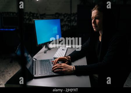 Side view of young hacker man wearing hooded stealing information, preparing to break password sitting at desk in dark room with brick wall. Stock Photo