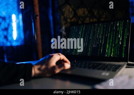 Close-up of unrecognizable hacker engaged in hacking into security system on laptop green binary code on screen with sitting at desk in dark basement Stock Photo