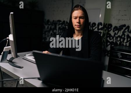 Front view of dangerous hacker man in sweatshirt typing code on laptop and desktop computer late night working and bypassing cyber security from dark Stock Photo
