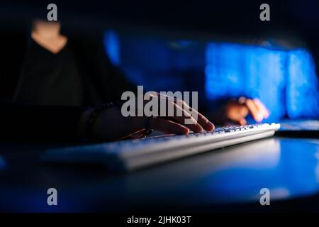 Close-up hands of unrecognizable hacker man working typing on keyboard laptop computer sitting at desk in dark room with blue neon lights. Stock Photo