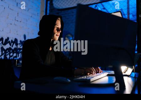 Low-angle view of wanted hacker coding virus ransomware using laptops and computers sitting at table in dark basement room with blue neon lights. Stock Photo