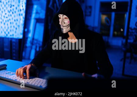 Wanted hacker man wearing in anonymous mask engaged in hacking into security systems, sitting in dark basement room with blue neon lights. Stock Photo