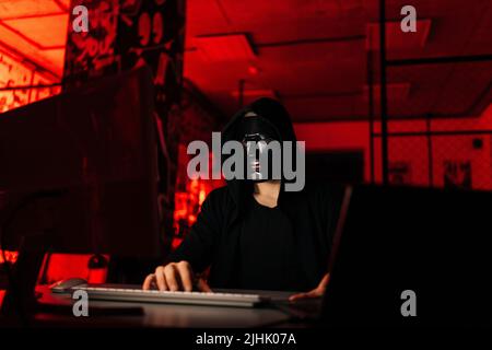 Dangerous unrecognizable hacker man wearing in black mask and sweatshirt hacking online web site using laptop and desktop Stock Photo