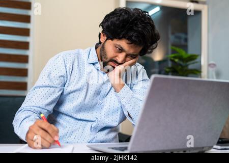 Tired young man while working on laptop by taking notes at office desk - concept of job frustration, routine work life and exhaustion. Stock Photo