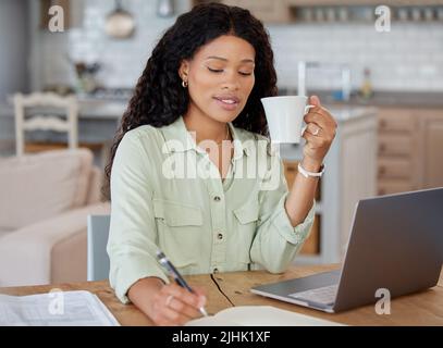 Working from home is so comfortable for me. a young woman writing notes and drinking coffee while working on a laptop at home. Stock Photo