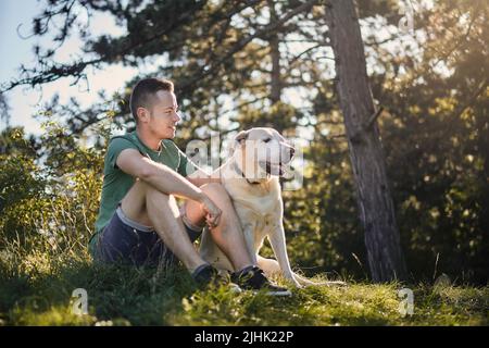 Man with his dog sitting in grass under tree. Pet owner enjoying trip with his labrador retriever during sunny summer day. Stock Photo