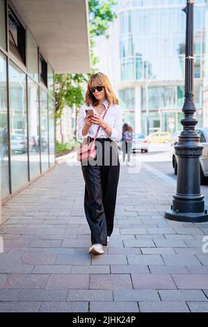 Attractive woman wearing sunglasses and text messaging while walking on sidewalk in the city. Stock Photo