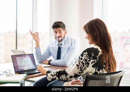 Teamwork at the office. Businesswoman and businessman sitting at desk and consulting about business plan. Wondering businessman raising his hand. Stock Photo