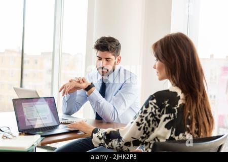 Teamwork at the office. Businesswoman and businessman sitting at desk and consulting about business plan. Busy professional man checking time on his w Stock Photo