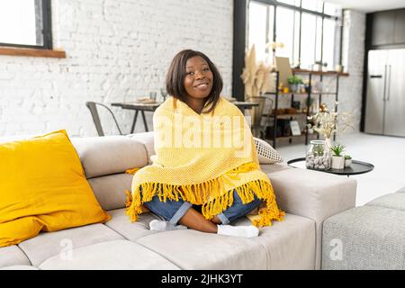 Serene African american woman enjoying weekend at home, sitting on the sofa wrapped with blanket and laughing, calm lady resting, looks at the camera and smiles Stock Photo