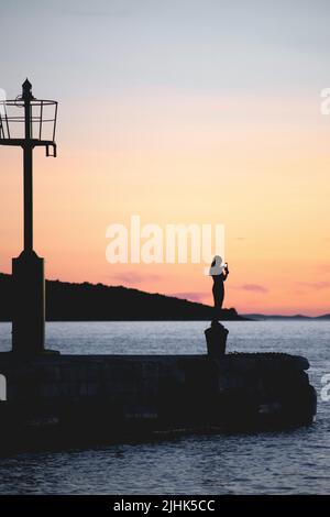 woman silhouette standing on ship mooring close to lighthouse holding phone and taking picture of beautiful sunset of the adriatic sea Stock Photo
