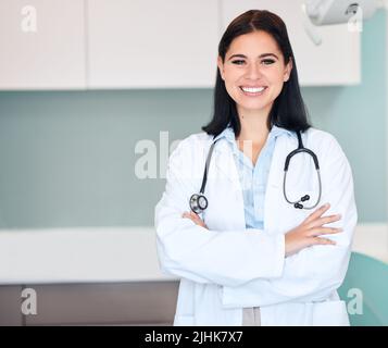 Confident young female dentist standing alone with her arms crossed in a medical office. One caucasian woman in a white coat with stethoscope. Oral Stock Photo