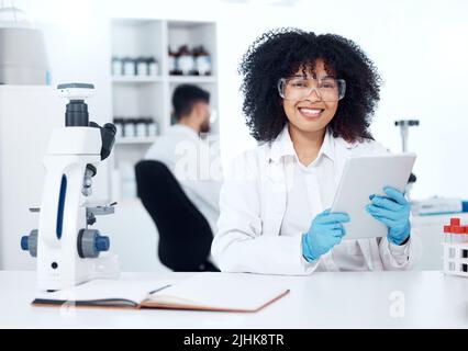 Portrait of young happy african american medical scientist with an afro using a digital tablet to record test results from a microscope. Mixed race Stock Photo
