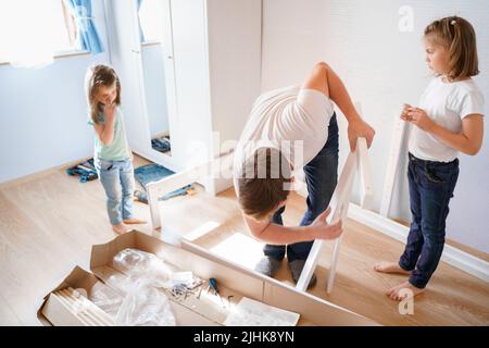 Dad,little girls daughters assemblle wooden baby bed according to instructions in kids room. Helping father, mounting children's furniture. Self-assem Stock Photo